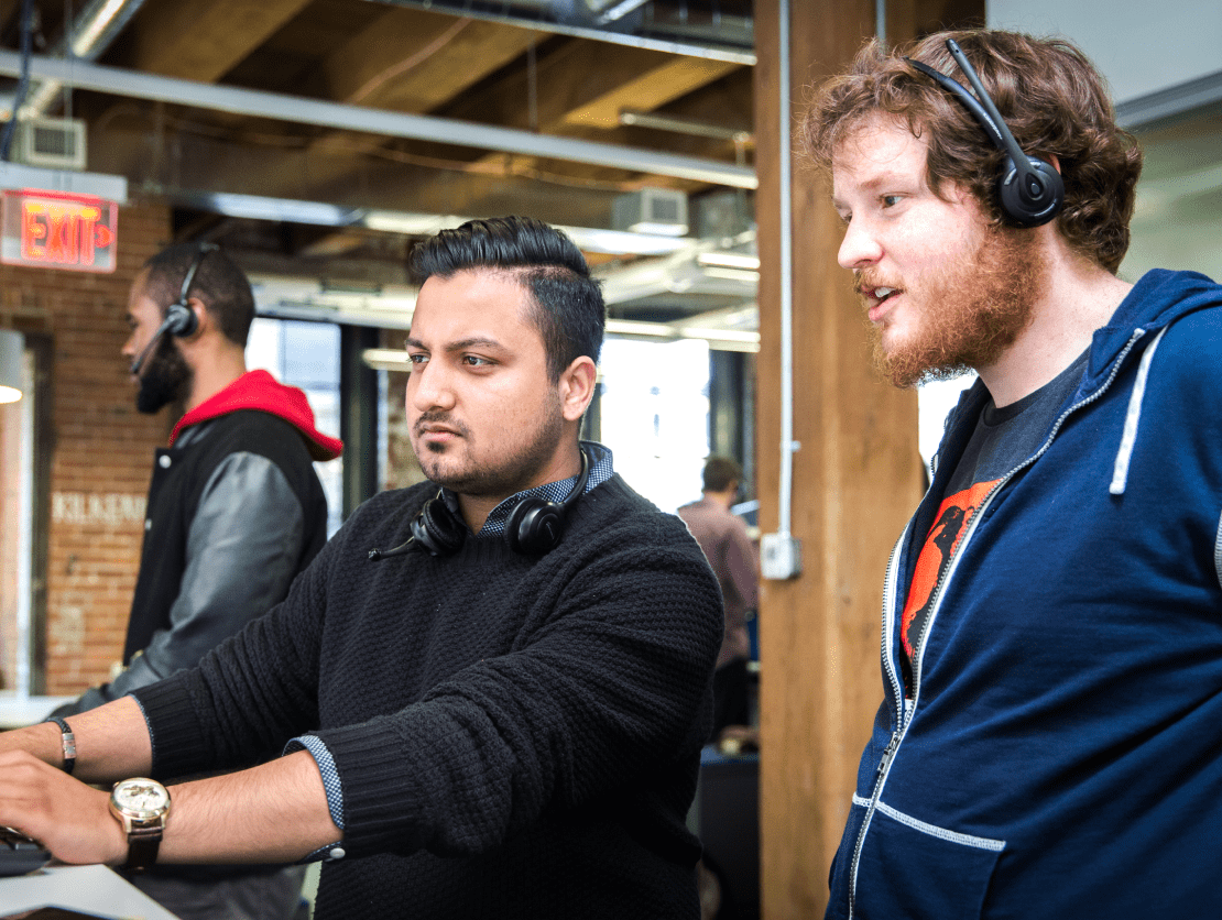 Two people wearing telecommunication headsets collaborating on a project on a singular computer.